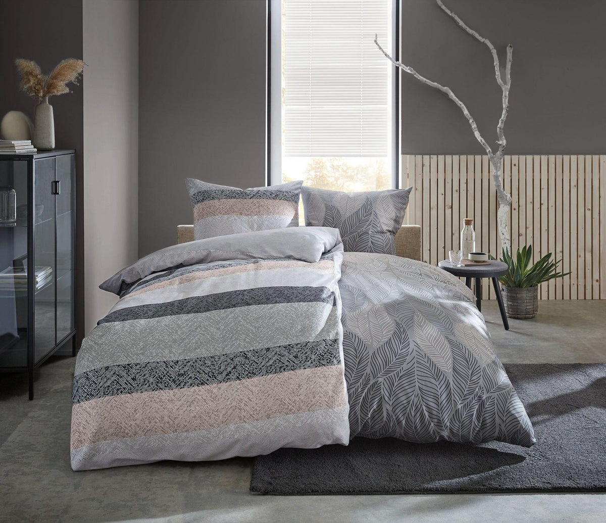 Graues Doppelbett mit gemusterter Bettwäsche vor grauer Wand und Fenster