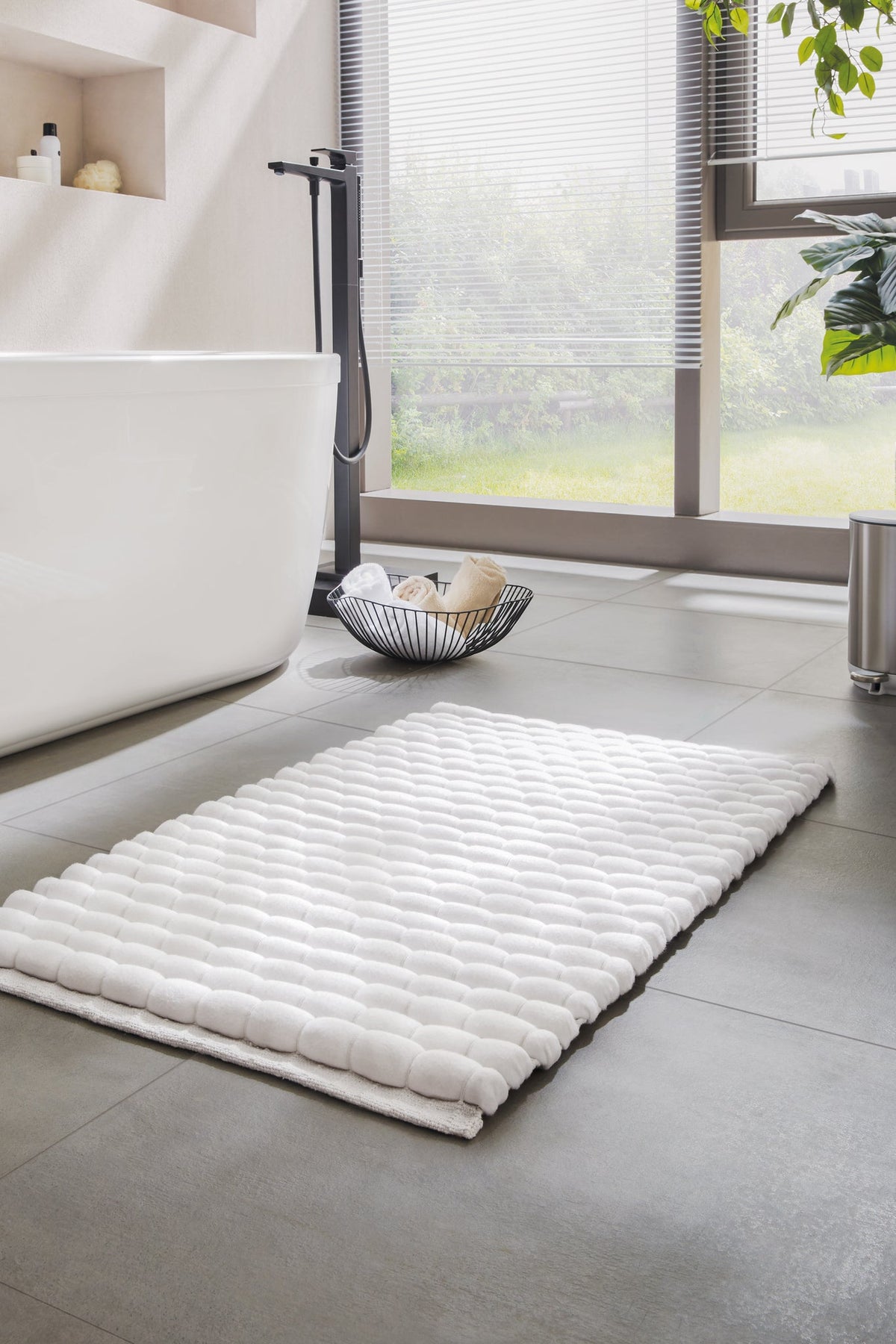 Weiße Badematte vor Badewanne auf grauen Fliesen im hellen Badezimmer