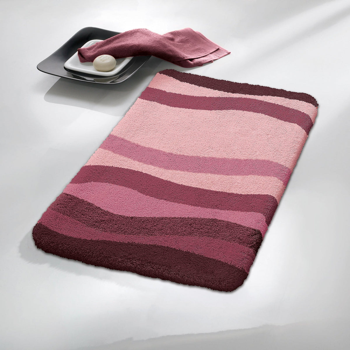 Badematte mit rosa und bordeauxfarbenen Wellen, Seife und Tuch daneben
