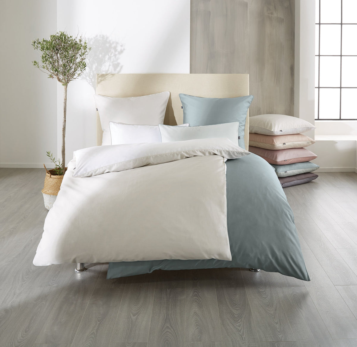 Doppelbett mit weiß-blauer Bettwäsche, Kissenstapel und Zimmerpflanze