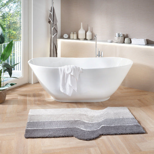 Freistehende Badewanne mit Handtuch und grauem Badteppich vor Fenster