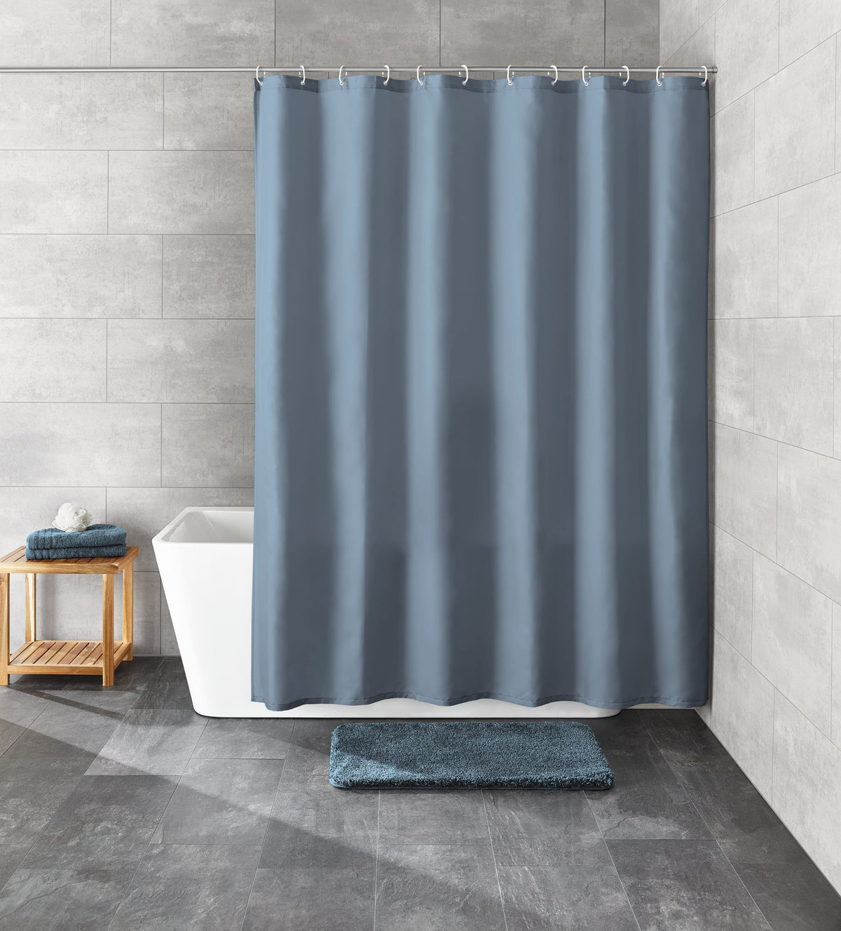 Grauer Duschvorhang vor Badewanne mit Holzhocker und blauer Badematte