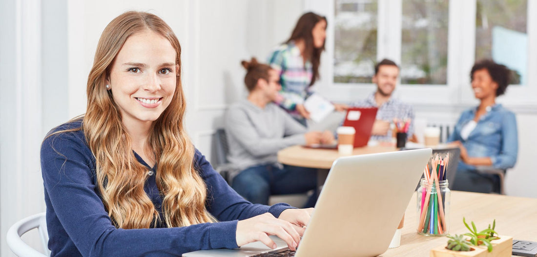 Junge Frau arbeitet lächelnd am Laptop, Kollegen im Hintergrund
