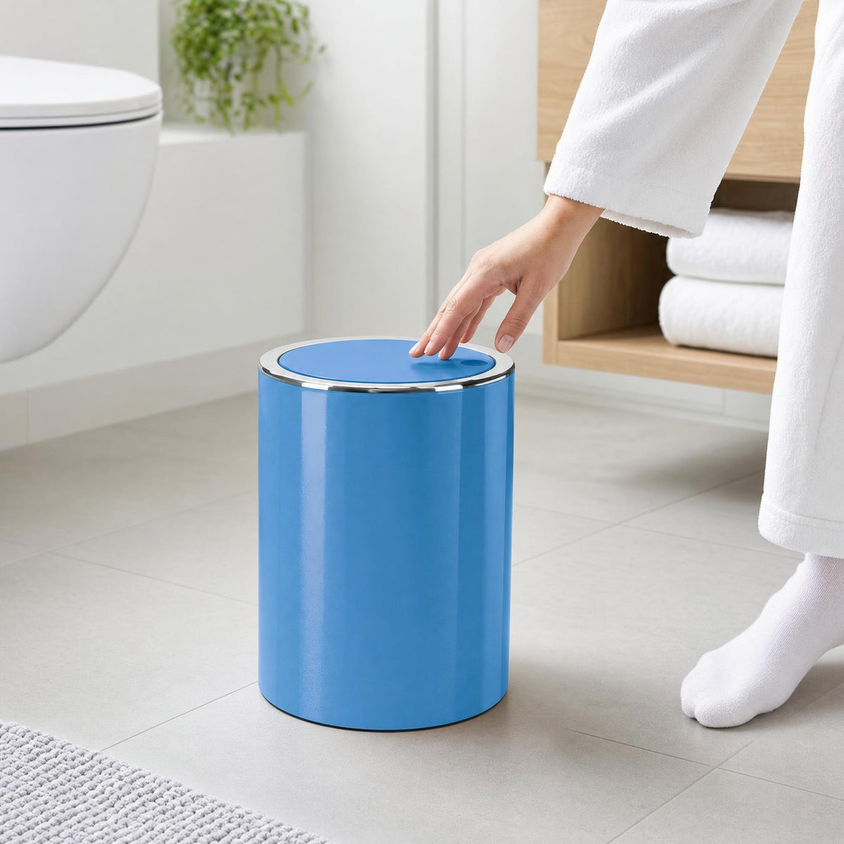 Person im Bademantel öffnet einen blauen Mülleimer im hellen Bad.