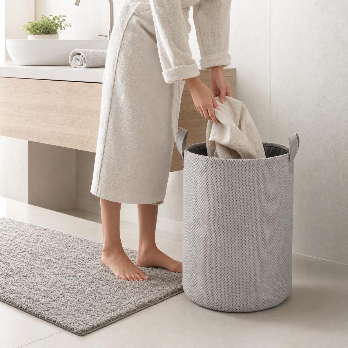 Ein grauer Wäschekorb steht im Bad, während ein Handtuch hineingelegt wird.