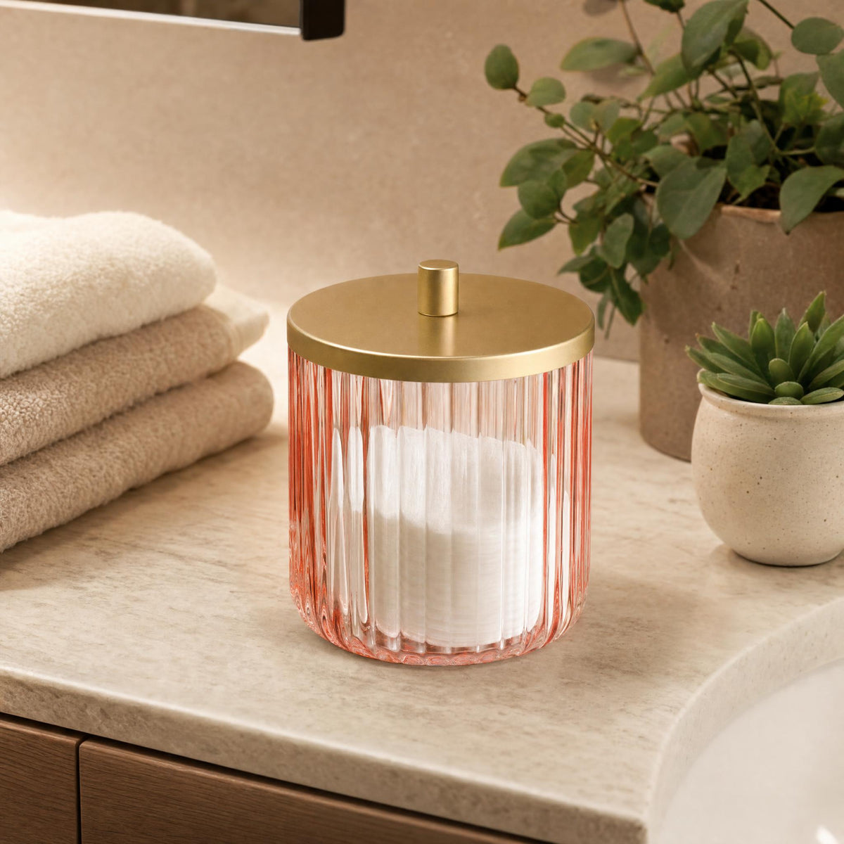 Rosa Glasdose mit Golddeckel auf Waschtisch neben Handtüchern und Pflanzen.