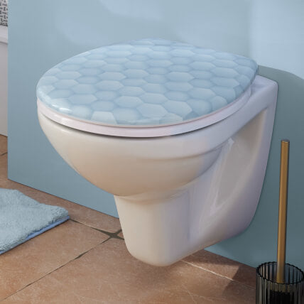 Wand-WC mit hellblauem WC-Sitz im Bad neben WC-Bürste und Badteppich.