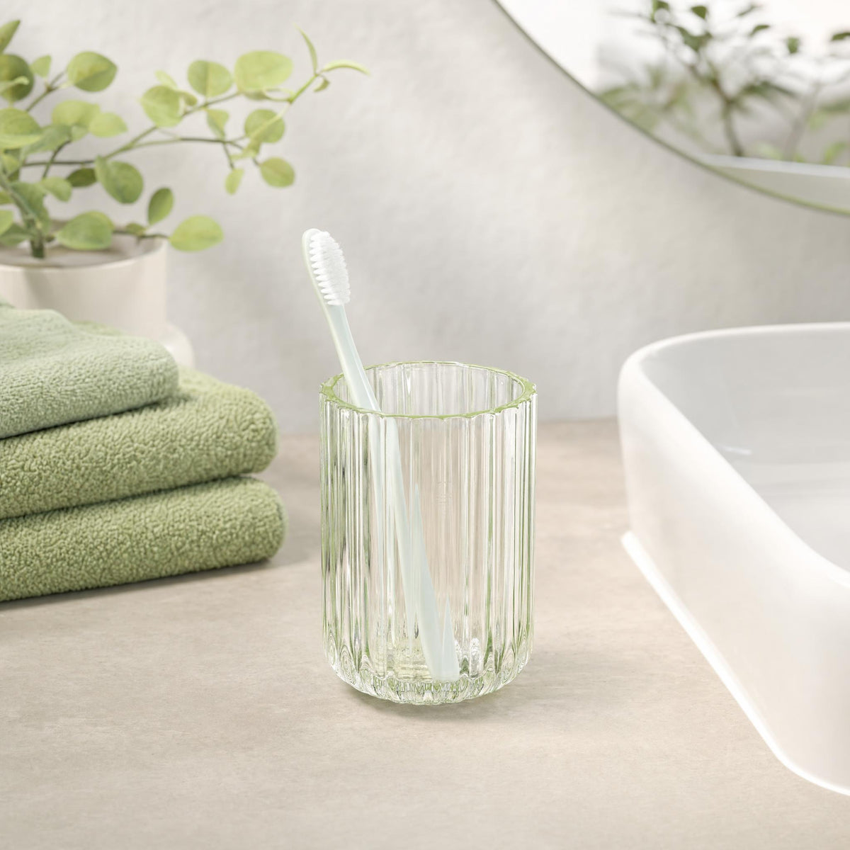Zahnbürste im geriffelten Glas neben Waschbecken und grünen Handtüchern.