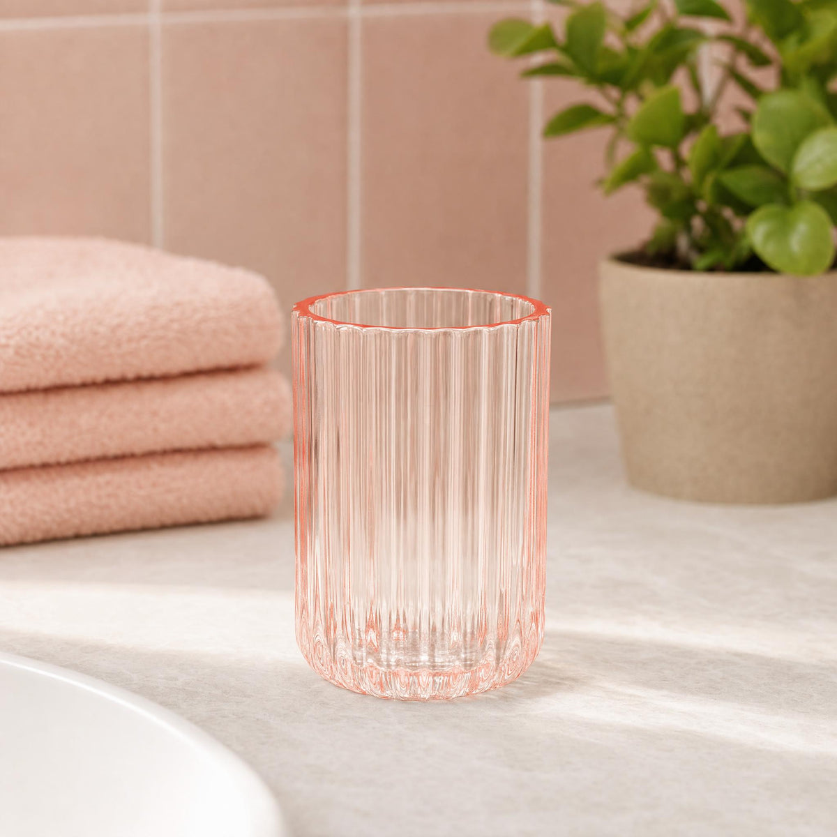 Geriffeltes rosa Glas auf Waschtisch neben Handtüchern und Pflanze.